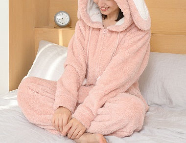 ピンク色のウサギ着ぐるみパジャマを着てフードをかぶりベッドに座る女性